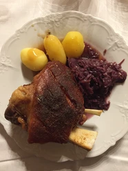 Gegrillte Schweinshaxe, serviert mit drei Salzkartoffeln und einer Portion Rotkohl auf einem weißen Teller.