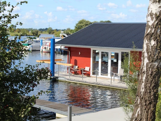 Ein rotes Holzhaus mit Terrasse liegt am Wasser, umgeben von Booten und Natur. Ein Birkenstamm im Vordergrund.