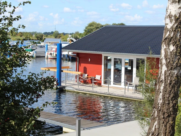 Wieltsee Smokey Island Ein rotes Holzhaus mit Terrasse liegt am Wasser, umgeben von Booten und Natur. Ein Birkenstamm im Vordergrund.