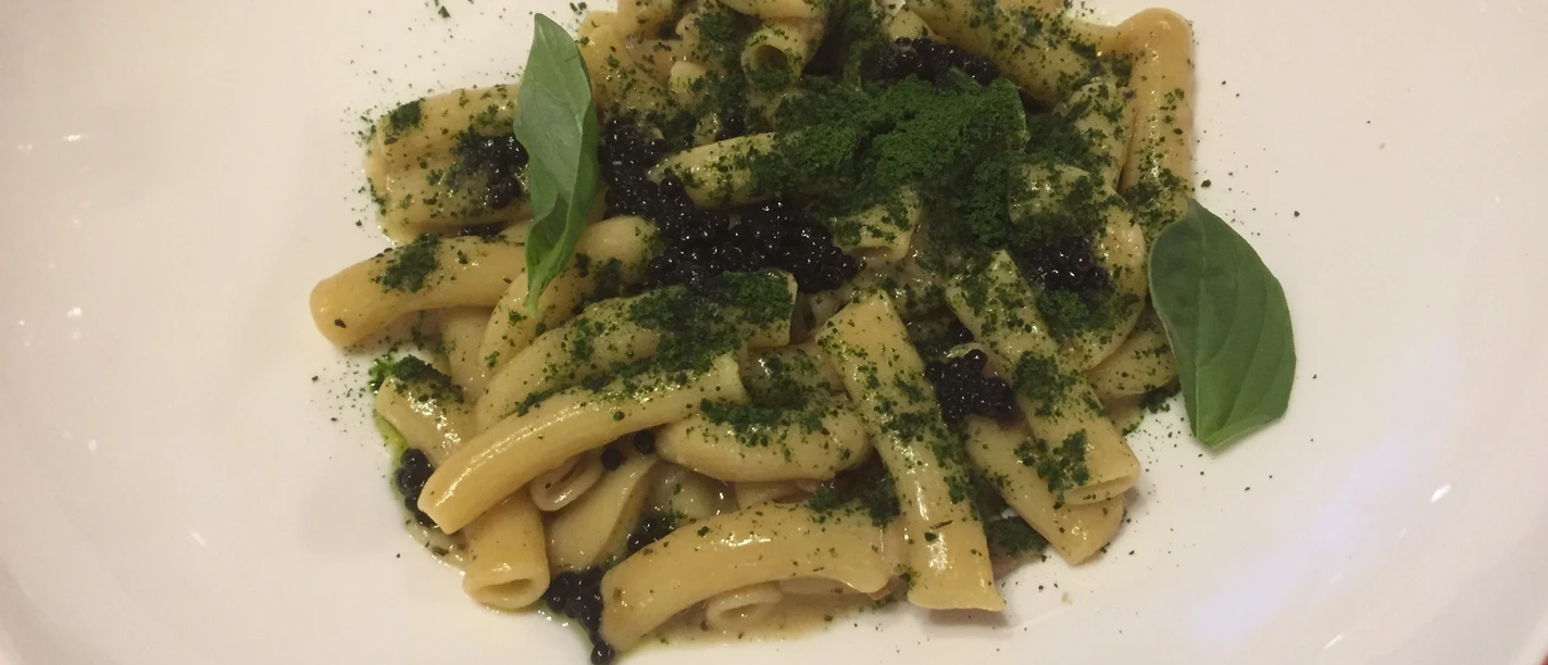 A white plate presents short tubular noodles garnished with fresh basil leaves and spices.