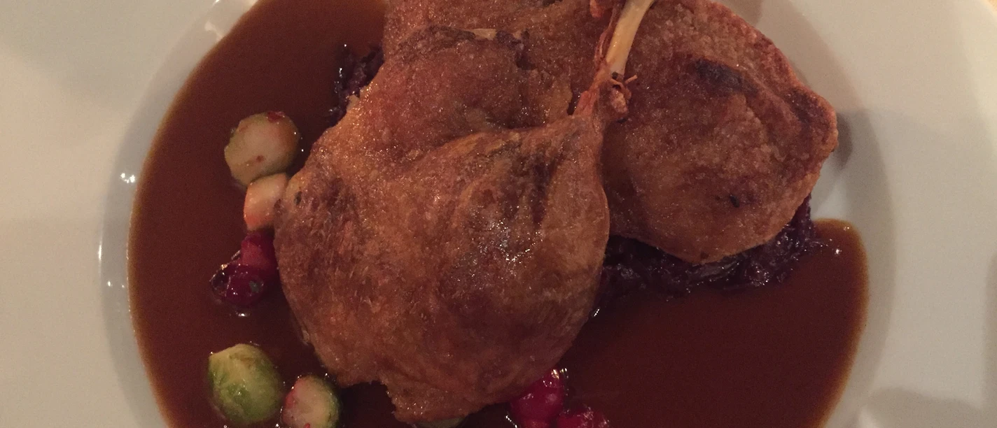 Dish of crispy goose leg served on a dark sauce, garnished with Brussels sprouts and cranberries.