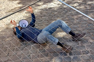 NO GO, Motjaba Izadizad Performer liegt hinter einem Metallgitter auf Kopfsteinpflaster, Megafon neben dem Kopf