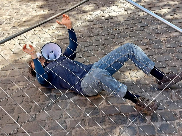 NO GO, Motjaba Izadizad Performer liegt hinter einem Metallgitter auf Kopfsteinpflaster, Megafon neben dem Kopf