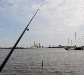 Angeln Emden.jpg Eine Angelrute im Vordergrund mit Blick auf den Emder Hafen, Schiffe und Kräne im Hintergrund.