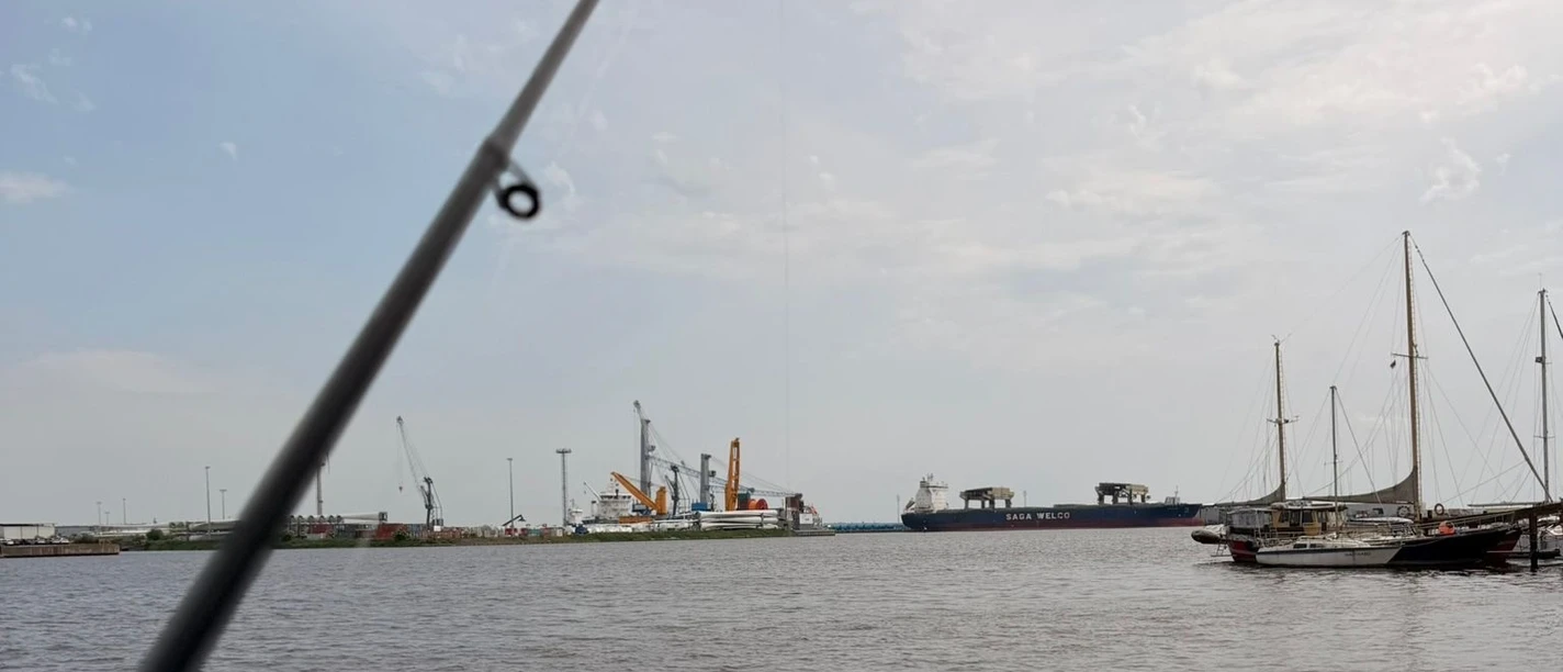 Angeln Emden.jpg Eine Angelrute im Vordergrund mit Blick auf den Emder Hafen, Schiffe und Kräne im Hintergrund.