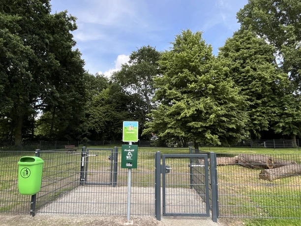 Hundefreilauffläche am Hahnentanger See Eingang zur umzäunten Hundewiese unter Bäumen mit Tor, Kotbeutelspender und Mülleimer.
