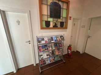 Toilette in der Tourist-Information Steinhude Das Bild zeiigt zwei Eingänge zu den Toiletten, links Damen und rechts das Herren WC. In der Mitte zwischen den Türen steht ein Aufsteller mit Flyern und darüber hängt ein dekoratives Fenster mit bunten Glasscheiben.