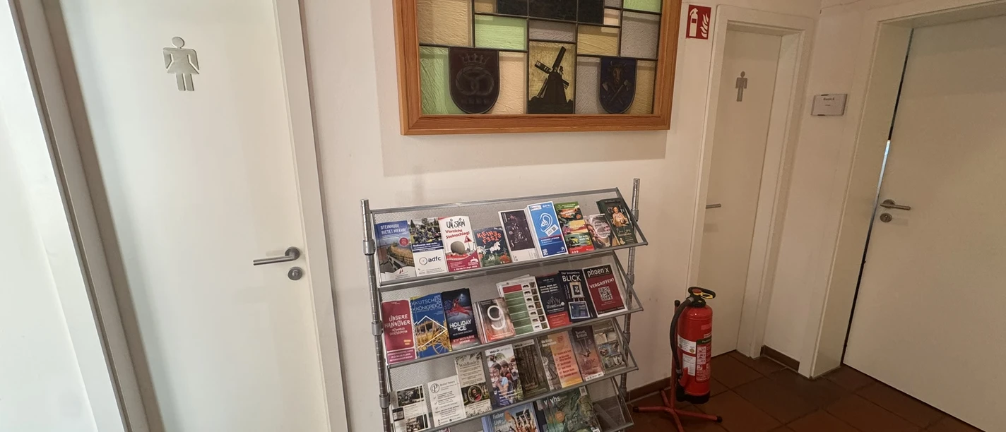 Toilette in der Tourist-Information Steinhude Das Bild zeiigt zwei Eingänge zu den Toiletten, links Damen und rechts das Herren WC. In der Mitte zwischen den Türen steht ein Aufsteller mit Flyern und darüber hängt ein dekoratives Fenster mit bunten Glasscheiben.