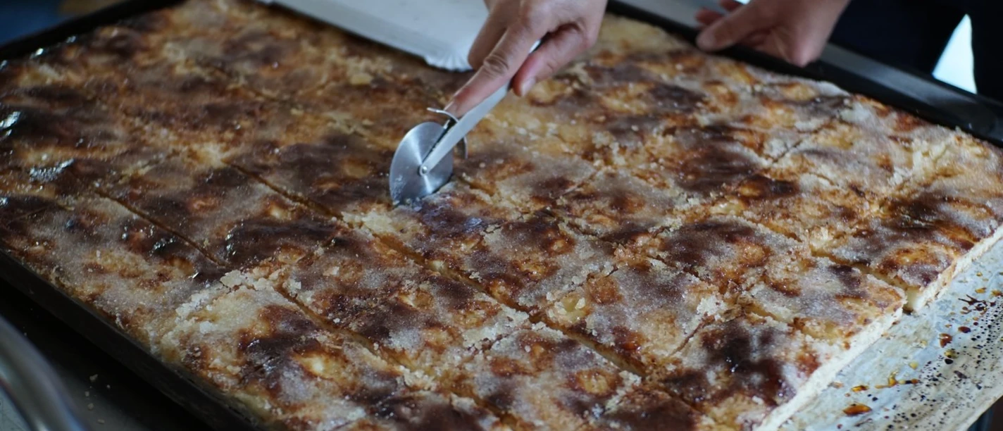 Backtag 2024.jpg Eine Hand schneidet mit einem Teigrad frischen Butterkuchen vom Blech in rechteckige Stücke.