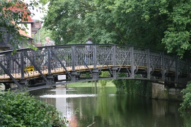 02-Herthabrücke.jpg Herthabrücke aus schwarzem Metall überspannt einen grünen Wasserlauf, umgeben von üppigen Bäumen.