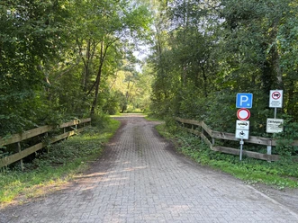 P11 Ein Steinweg führt in Richtung Parkplätze. Rechts und Links stehen halbhohe Holzzäune und rechtsseitig sind einige Hinweisschilder zu sehen.