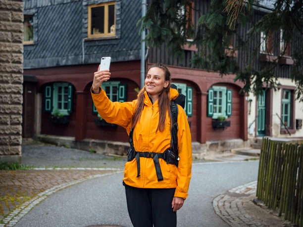 Frau in gelber Jacke macht ein Selfie mit ihrem Smartphone in einer malerischen Straße.