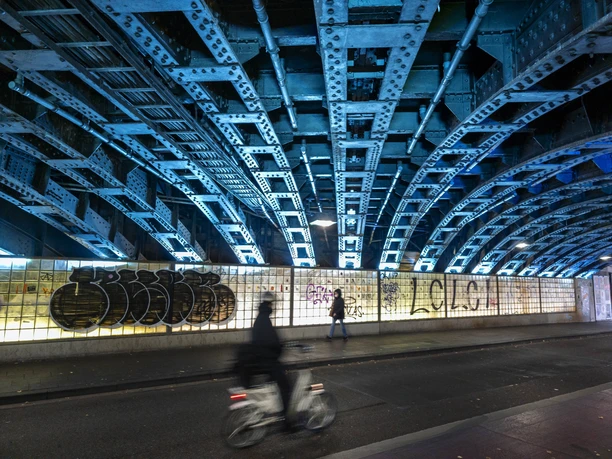 Eigelstein Tunnel Blue-lit steel beams at Eigelstein, featuring a graffiti wall, cyclists, and passersby