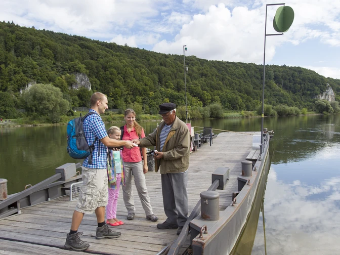 Auf der Gierseilfähre in Matting Vier Personen stehen auf einer Gierseilfähre über einem Fluss in einer grünen Landschaft.
