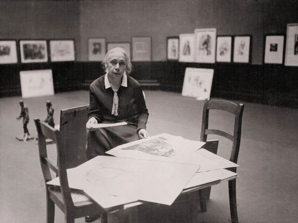 Käthe Kollwitz in der Preußischen Akademie der Künste, Berlin 1927 Historische Aufnahme von Käthe Kollwitz im Ausstellungsraum an einem Tisch mit Grafiken.