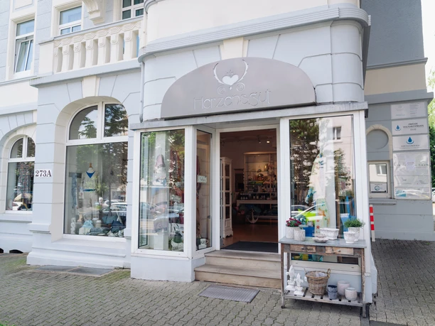 Herzensgut_Bad Wilhelmshöhe Der Eingang von dem Geschäft Herzengut in Wilhelmshöhe Kassel ist abgebildet. Das Gebäude ist grau & weiß, über der Tür hängt ein Schild mit der Beschriftung "Herzensgut".
