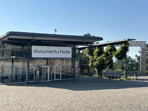 Das Gebäude der documenta Halle mit Flachdach, Glasfront und einem Schild mit der Aufschrift "documenta Halle" ist sichtbar. Im Hintergrund befindet sich das documenta Außenkunstwerk "Rahmenbau". Vor dem Gebäude ist der Platz gepflastert.
