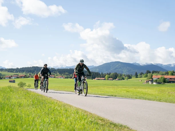 Radgruppe fährt auf Landstraße durch grüne Wiesen vor Alpenkulisse bei Bad Bayersoien