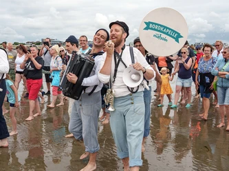 Die BORDKAPELLE von mahoin beim Wattenlaufen mit Musik Zwei Männer mit Instrumenten im Watt. Hinter ihnen mehrere Menschen Meschen.