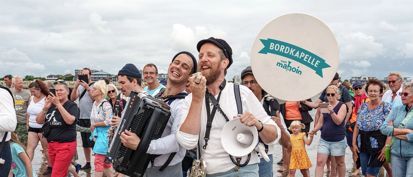 Die BORDKAPELLE von mahoin beim Wattenlaufen mit Musik Zwei Männer mit Instrumenten im Watt. Hinter ihnen mehrere Menschen Meschen.