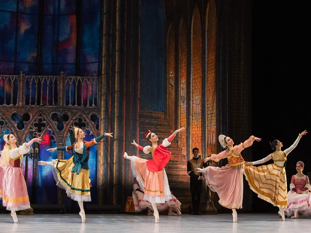 Schwanensee- GRAND CLASSIC BALLET Mehrere Ballerinen tanzen in farbigen Kostümen vor gotischer Bühnenkulisse