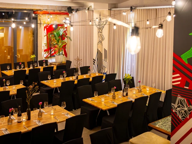 Alte Textilfabrik - Veranstaltungsraum der Alten Textilfabrik Blick in einen eingedeckten Gastronomie- oder Veranstaltungsraum der Alten Textilfabrik mit Holztischen, schwarzen Stühlen, Lichterketten und industriellem Interieur. Raumteiler und Wandgestaltung setzen farbige Akzente.