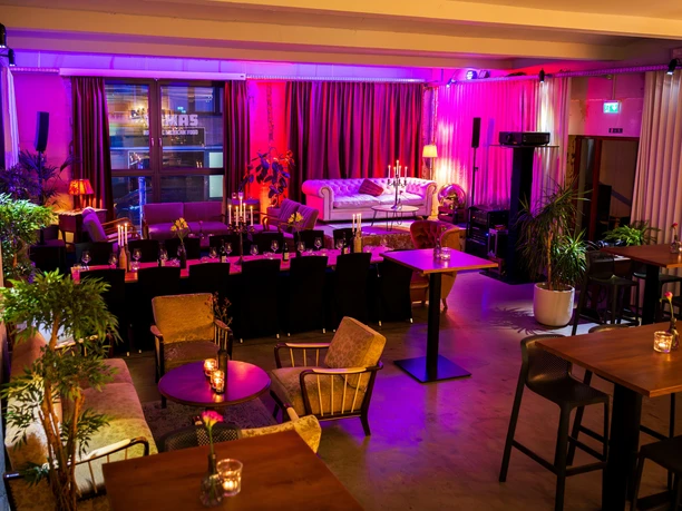 Old Textile Factory - Lounge and Event Area A lounge and event area in the Old Textile Factory featuring sofas, armchairs, candles, plants, speakers, and colorful lighting. In the foreground are tables set for guests.