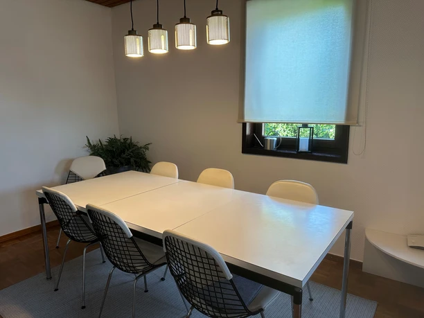 Esszimmer des Ferienhauses am Ritzberg Weißer Esstisch mit sieben Stühlen, Pendelleuchte, im Hintergrund Fenster mit Rollo