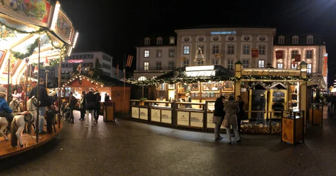 7 Schwaben auf dem Kasseler Märchenweihnachtsmarkt Der beleuchtete Stand der 7 Schwaben ist im Hintergrund sichtbar. Zwei Besucher stehen vor der Hütte. Links ist auf dem Bild noch ein Karussell erkennbar.