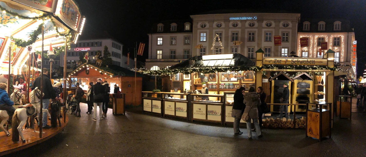 7 Schwaben auf dem Kasseler Märchenweihnachtsmarkt Der beleuchtete Stand der 7 Schwaben ist im Hintergrund sichtbar. Zwei Besucher stehen vor der Hütte. Links ist auf dem Bild noch ein Karussell erkennbar.