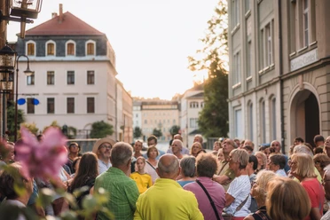 Nachtwächterführung-11.jpg Eine Gruppe von Menschen steht in einer historischen Altstadtstraße und hört aufmerksam einer Stadtführung zu, während die Abendsonne die Gebäude warm beleuchtet.