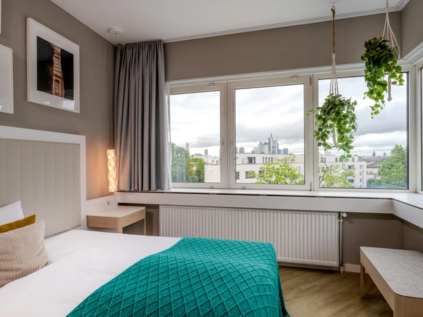 The Blasky Hotel Frankfurt ROOFTOP-Modernes Hotelzimmer. Modernes Hotelzimmer mit großem Fenster, Blick auf die Frankfurter Skyline, einem Bett mit grüner Decke und hängenden Zimmerpflanzen.