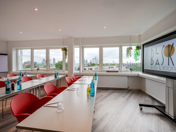 The Blasky Hotel Frankfurt ROOFTOP- Heller Tagungsraum. Heller Tagungsraum mit Tischen und roten Stühlen, einem großen Präsentationsbildschirm und einer Fensterfront mit Blick auf die Frankfurter Skyline.