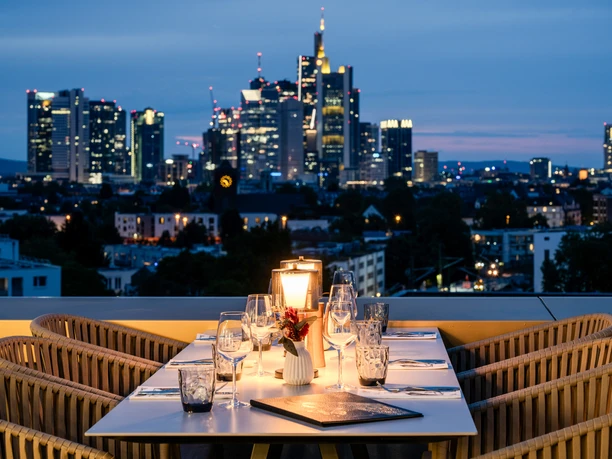 Vienna House by Wyndham Frankfurt South —An evening view from the hotel's rooftop terrace An evening view from a hotel rooftop terrace, with a table set, overlooking the illuminated Frankfurt skyline at dusk.