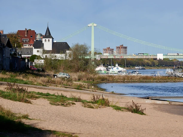 The banks of the Rhine in Rodenkirchen Sandy beach on the Rhine in Rodenkirchen with a church, boats, a pier, and the Rodenkirchen Bridge