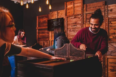 Gamer-Sling Zwei Personen spielen an einem hölzernen Spielautomaten einen Puck sich gegenseitig zu. Im Hintergrund spielen weitere Personen. Es befinden sich mehrere Holzboxen an der Wand im Hintergrund.