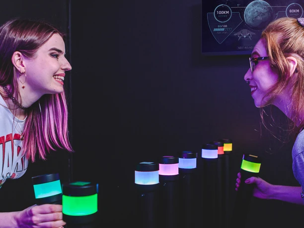 Gamer-Square Zwei Frauen stehen sich gegenüber, in der Mitte stehen unterschiedlich farbig beleuchtete Pfosten. Beide Personen halten je einen Pfosten in der Hand.
