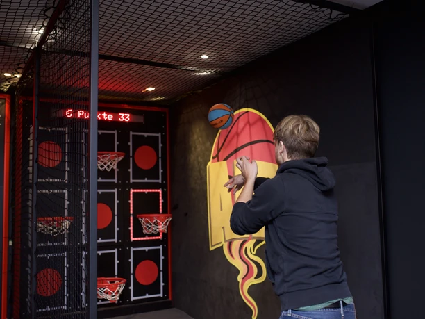 Gamer-Air An der Wand befinden sich Basketballkörbe und Vierecke mit einem roten Punkt in der Mitte. Davor steht ein Mann der einen Ball in die Richtung der Wand wirft.