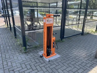Orange Fahrrad-Reparaturstation vor einem verglasten Fahrradunterstand am Campus.