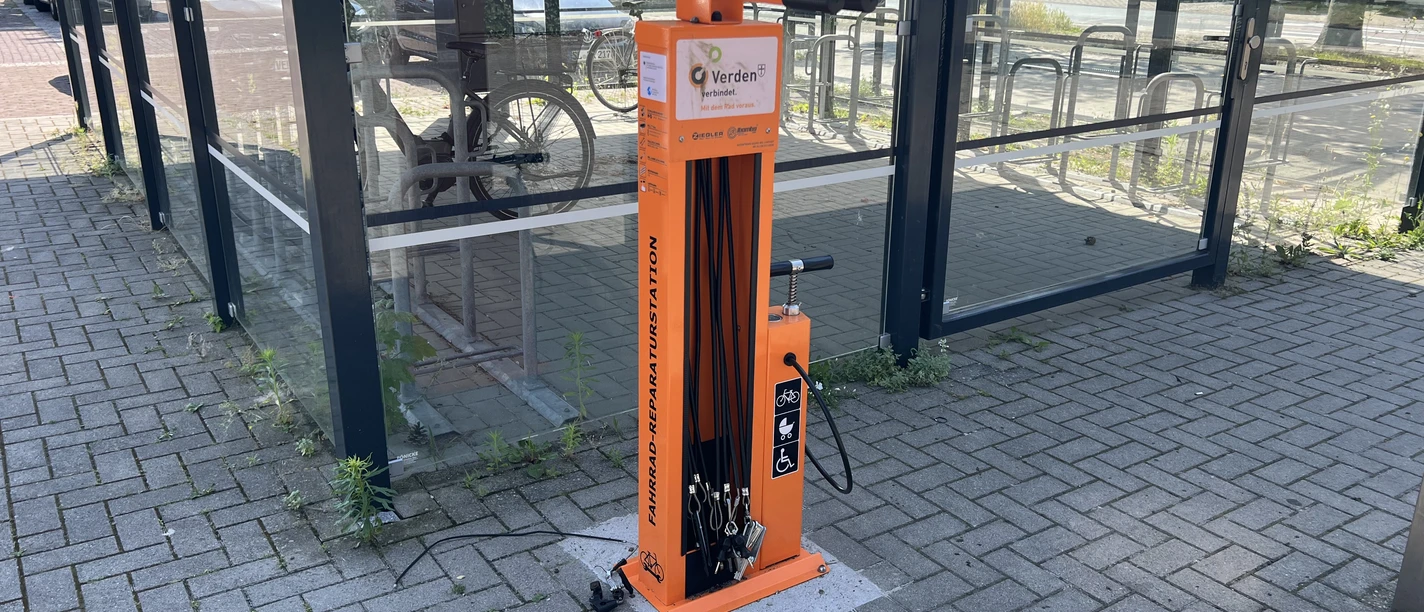 Fahrradstation Verdener Campus-Oberschule (5).jpeg Orange Fahrrad-Reparaturstation vor einem verglasten Fahrradunterstand am Campus.