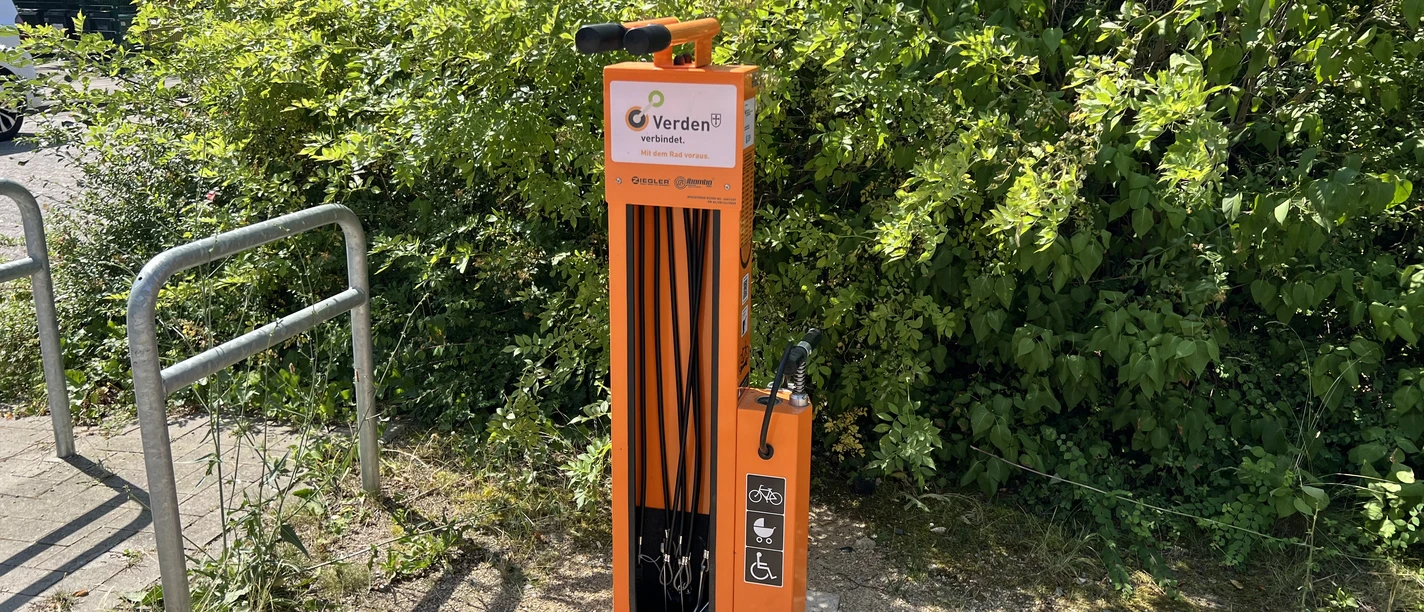Fahrradstation Oderplatz Verden (1).jpeg Orange Fahrradreparaturstation mit Werkzeugen vor grünen Büschen.
