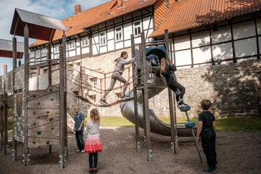 Kinder klettern auf einem Spielplatz vor Fachwerkhäusern und alter Steinmauer.