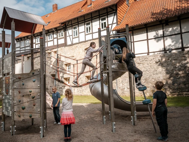 Kinder klettern auf einem Spielplatz vor Fachwerkhäusern und alter Steinmauer.