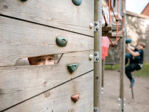 Ein Kind blickt durch eine Holzwand, ein anderes klettert im Hintergrund am Spielgerät.