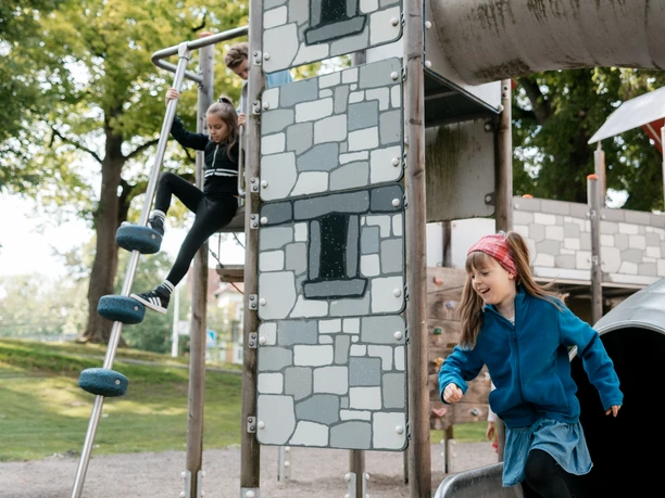 Kinder spielen auf einem Kletterturm mit Rutsche unter Bäumen.
