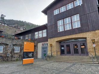 Eingang des Museums Rammelsberg mit orangefarbener Infotafel und historischem Gebäude.The entrance to the Rammelsberg Museum, featuring an orange information board and a historic building.