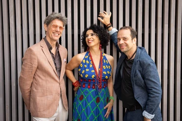 Bossarenova Trio The Bossarenova Trio poses smiling in front of vertical slats; the singer is wearing a colorful dress