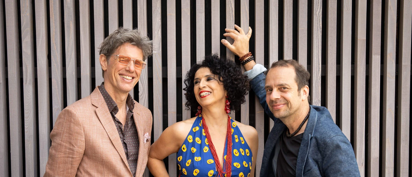 Bossarenova Trio The Bossarenova Trio poses smiling in front of vertical slats; the singer is wearing a colorful dress