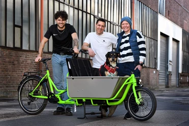 ccb colognecargo.bike Team Drei Personen mit Hund an grünem Lastenrad vor Kölner Backsteinfassade
