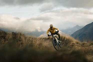 Conduite dynamique dans les virages en terrain alpin Sur le Flowtrail du col de Wurzenbord, un vététiste négocie avec dynamisme un virage serré, entouré de prairies alpines et de nuages spectaculaires.